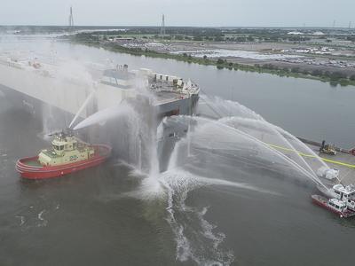 Tugboat spraying water on a docked car carrier during port firefighting operations